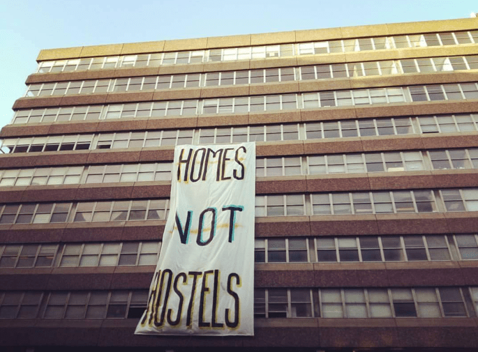 Urbanität steht in Bezug zum Treiben der politischen Stadt. Im Bild ein Plattenbau. Auf einem Banner steht "Homes not Hostels". Ein Aufruf zu mehr Lebensraum.