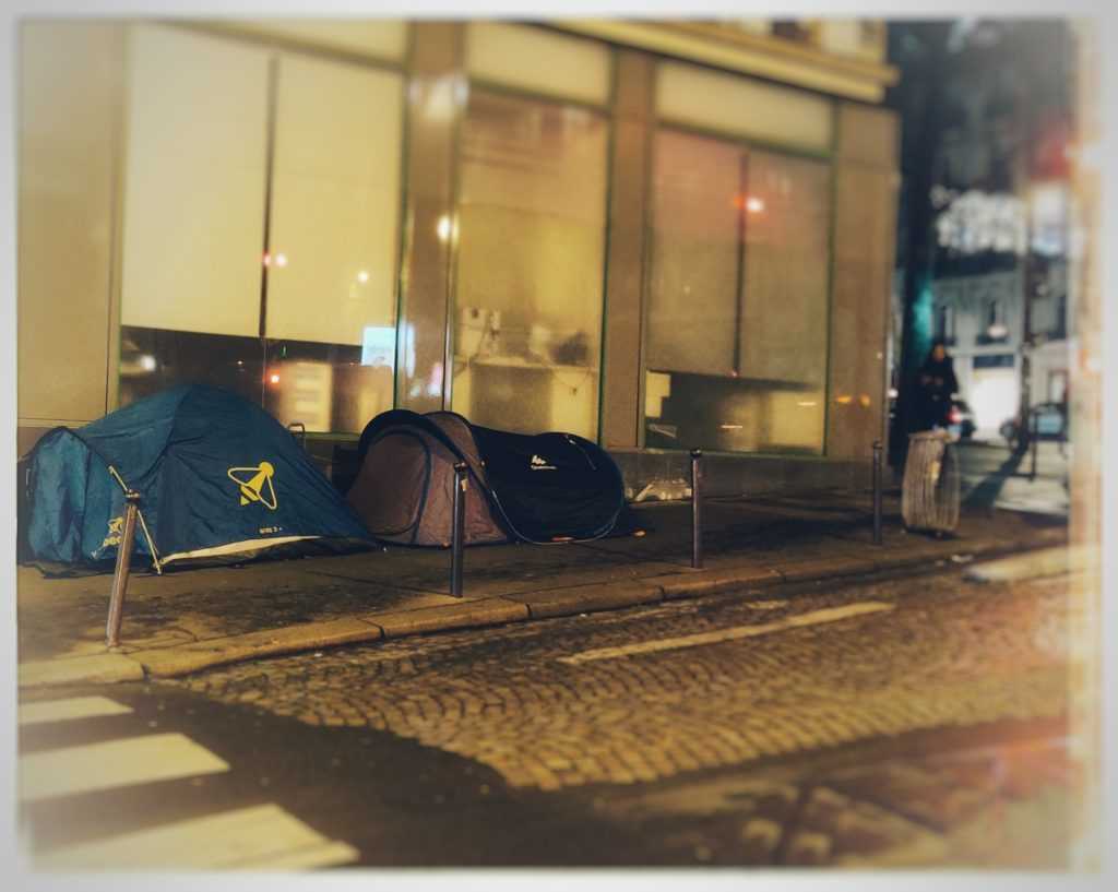 Die politische Stadt im Kontext von Armut und Obdachlosigkeit. Zwei Zelte von Obdachlosen in Paris. Die Autofahrbahn besteht aus Pflastersteinen, während das an die Zelte angrenzende Gebäude eine glatte Marmorfassade aufweicht. Das Bild wurde bearbeitet sodass der Licht und Schärfe Fokus auf die Zelte liegt. Die restlichen Farben, darunter das rot einer Ampel, werden verschwommen dargestellt. Das Photo wurde von Urbanauth 2020 in Paris aufgenommen.