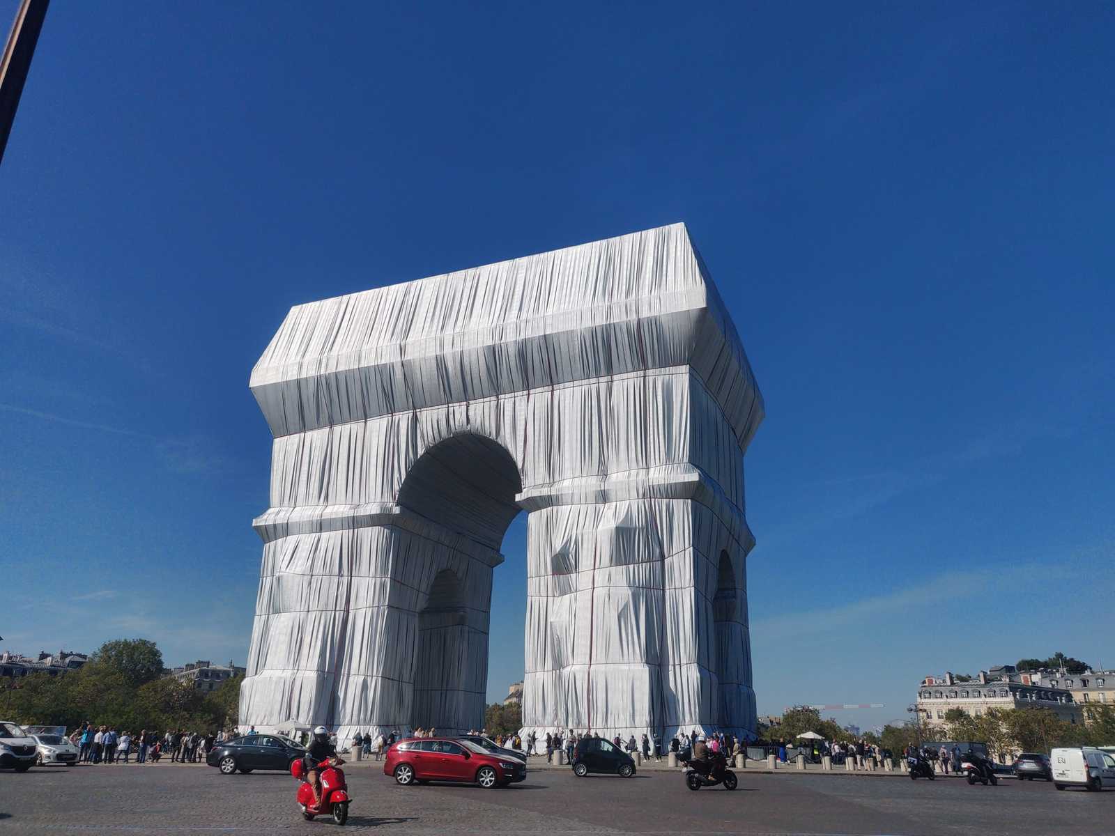 Paris, der verhüllte Triomphbogen vom Arc de Triomphe. Blauer Himmel. Viel Autoverkehr. Verhüllungskunst von Christo und Jeanne-Claude in einem posthumen Projekt. Das Gewebe zum verhüllen scheint silber und hinterlässt Falten.